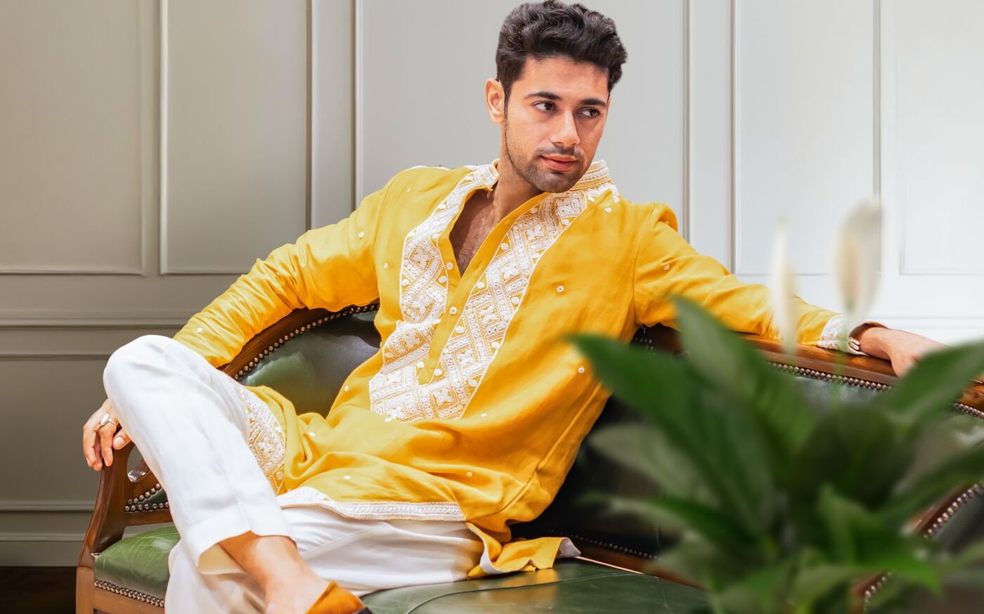 Man in a bright yellow embroidered kurta paired with white churidar pants, relaxing on a classic green vintage sofa in a modern interior setting